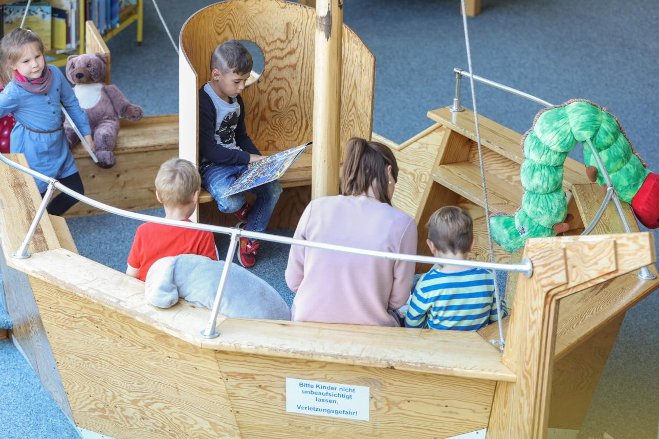 Lesen und Arbeiten in der Stadtbibliothek. Foto: Andrea Fabry Mehrere Kinder und eine Mutter sitzen auf einer Holzbank, die einem Boot nachempfunden ist und lesen Bücher.